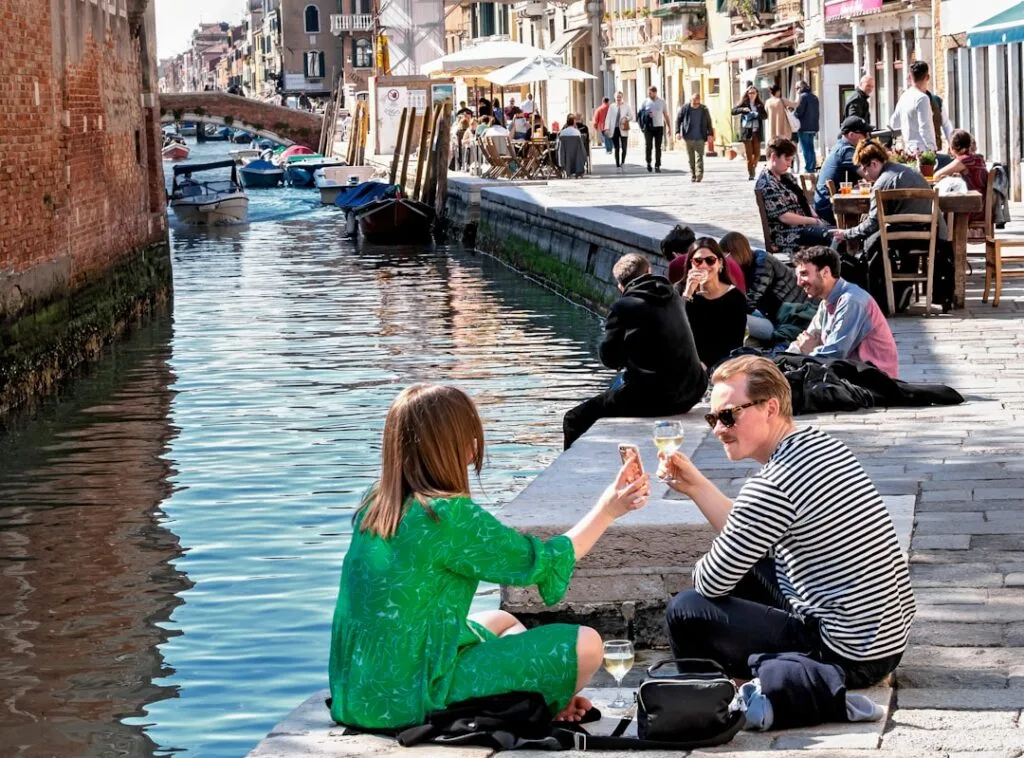 Couple trinquant au bord d'un canal, passants et bateaux en arrière-plan