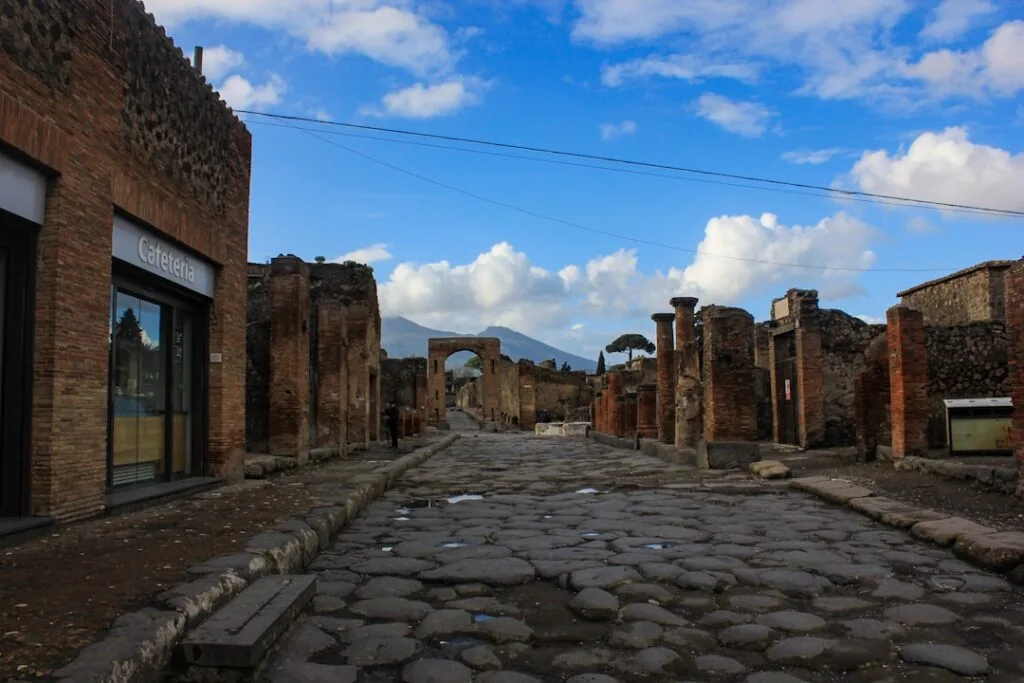 Wide cobbled Pompeii street flanked by ruins and distant Mount Vesuvius.