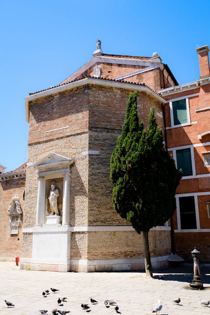 Backstein-Kapelle mit Statue und Zypresse auf sonniger Piazza.