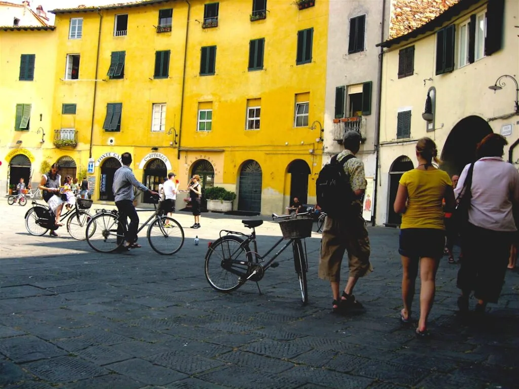 People and bicycles gathered in Lucca square in front of yellow buildings.