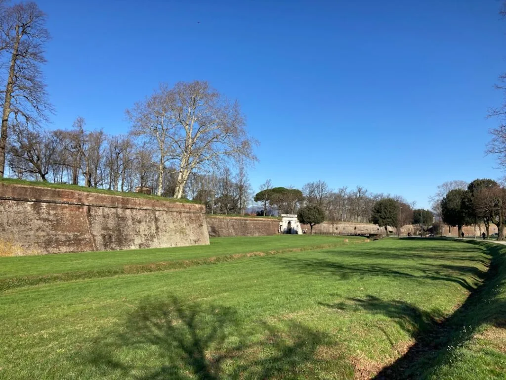 Grassy moat beside Lucca's stone walls and distant arched gate.