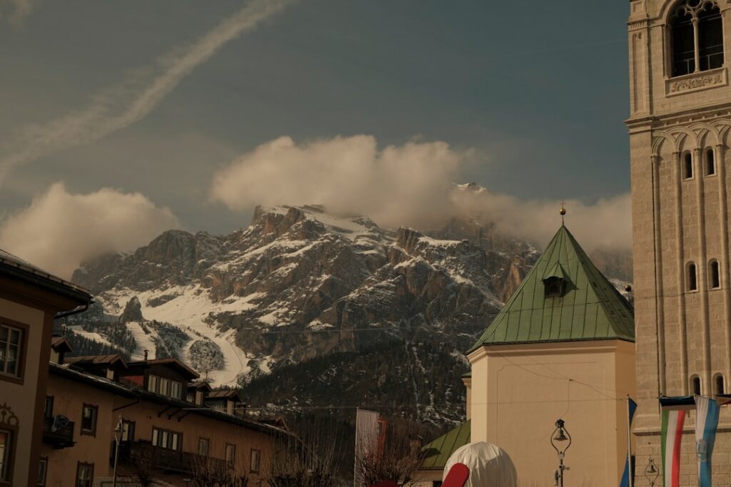 Bergdorf mit Kirchturm vor schneebedeckten Dolomiten-Gipfeln und Wolken