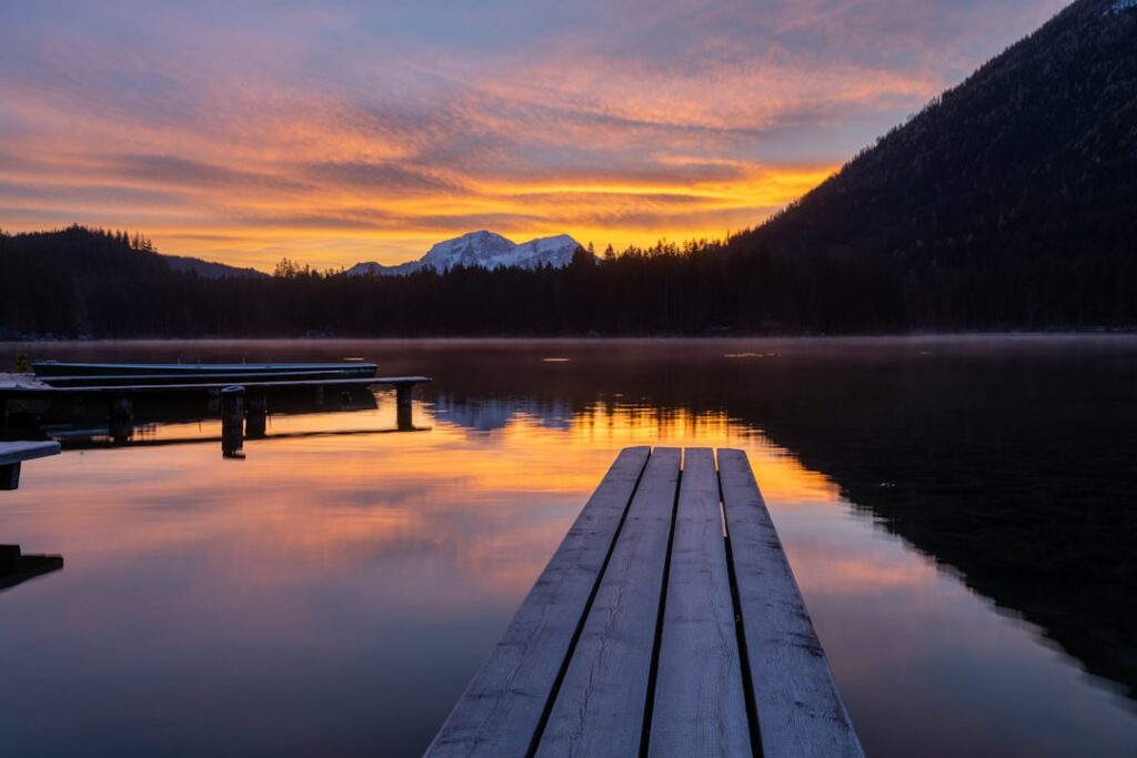 Holzsteg am Seeufer bei Sonnenaufgang, bunte Reflexe und Dolomiten-Silhouette
