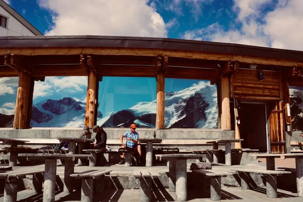 Terrasse en bois avec bancs et grandes fenêtres reflétant glaciers des Dolomites, visiteurs assis