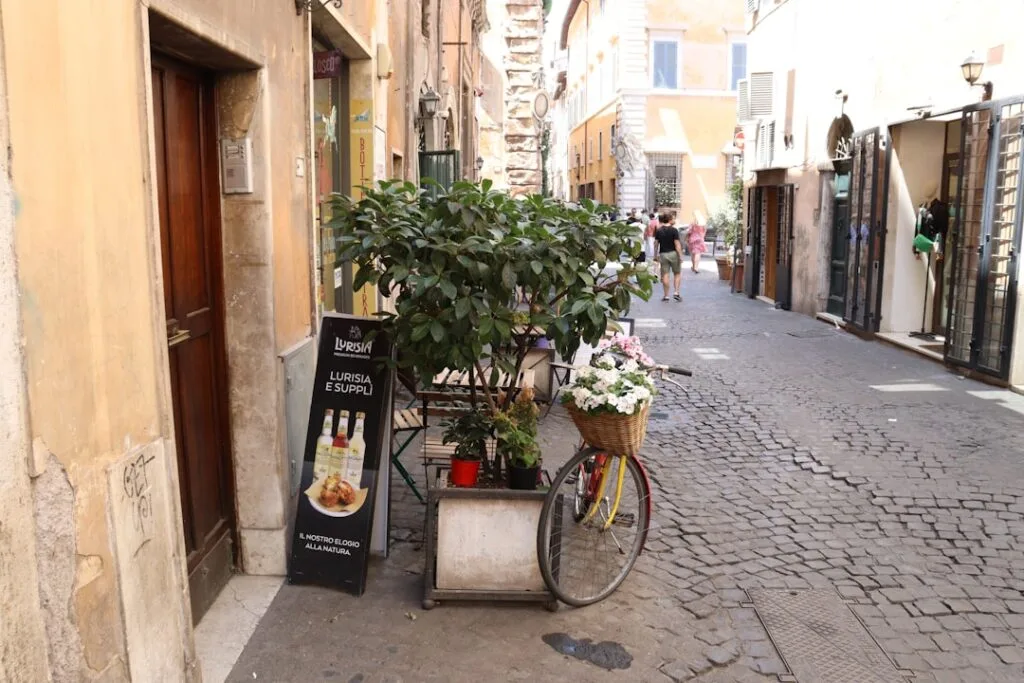 Rue pavée romaine avec bicyclette fleurie et panneau 'Lurisia e Supplì'