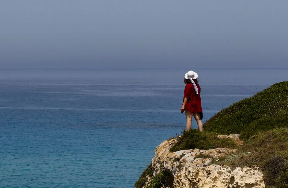 Woman in white hat stands on rocky cliff overlooking sea near Lecce