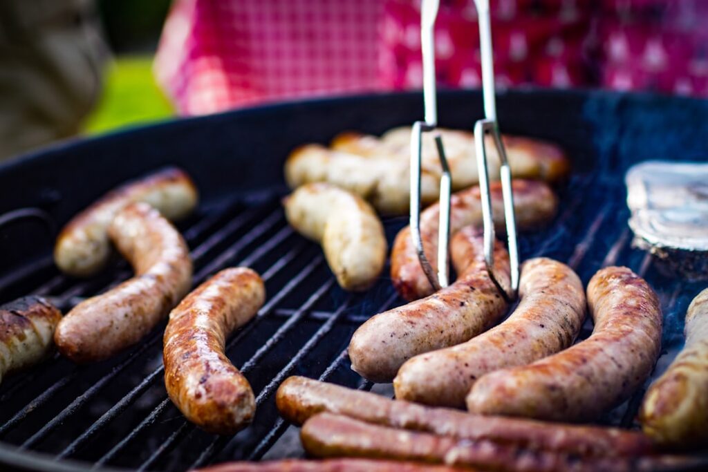 Bratwürste liegen auf Grillrost, Zange wendet eine Wurst