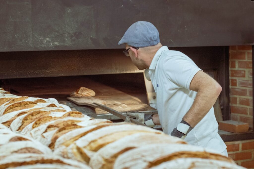 Bäcker schiebt Brot in traditionellen Ofen, Reihen frischer Laibe sichtbar