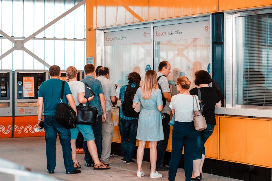 Rij mensen wachtend bij geel-oranje ticketloket, ruggen richting camera