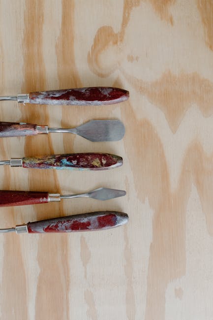 Four paint-splattered palette knives aligned on pale wooden plywood surface