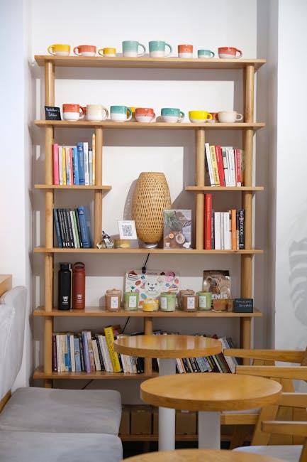 Wooden shelf of colorful mugs and books above small tables.