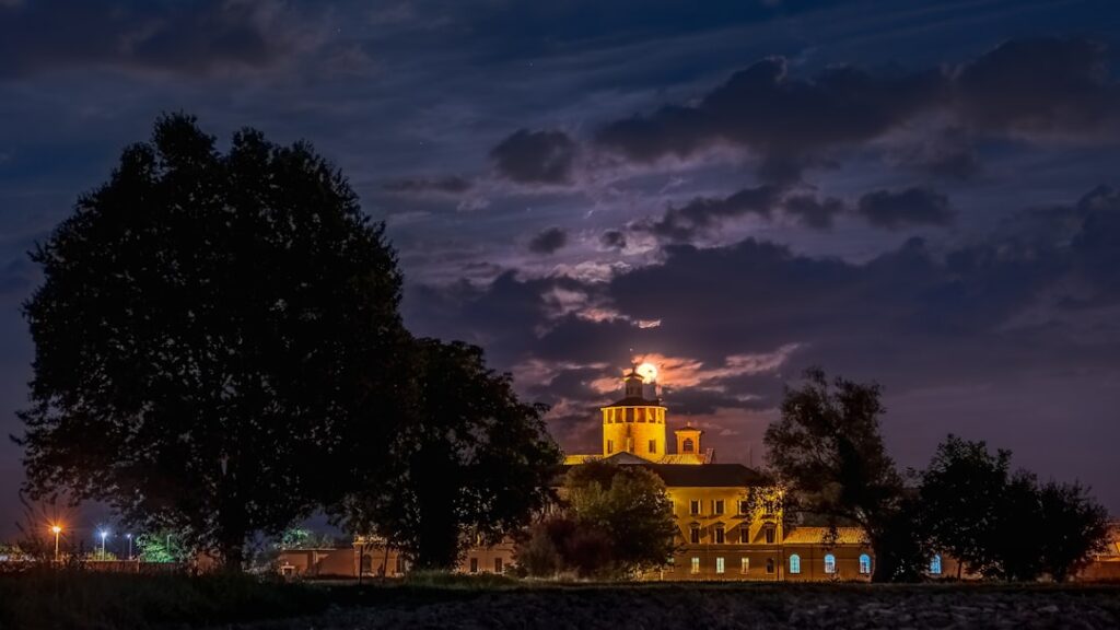 Illuminated historic building in Parma framed by trees under moonlit clouds