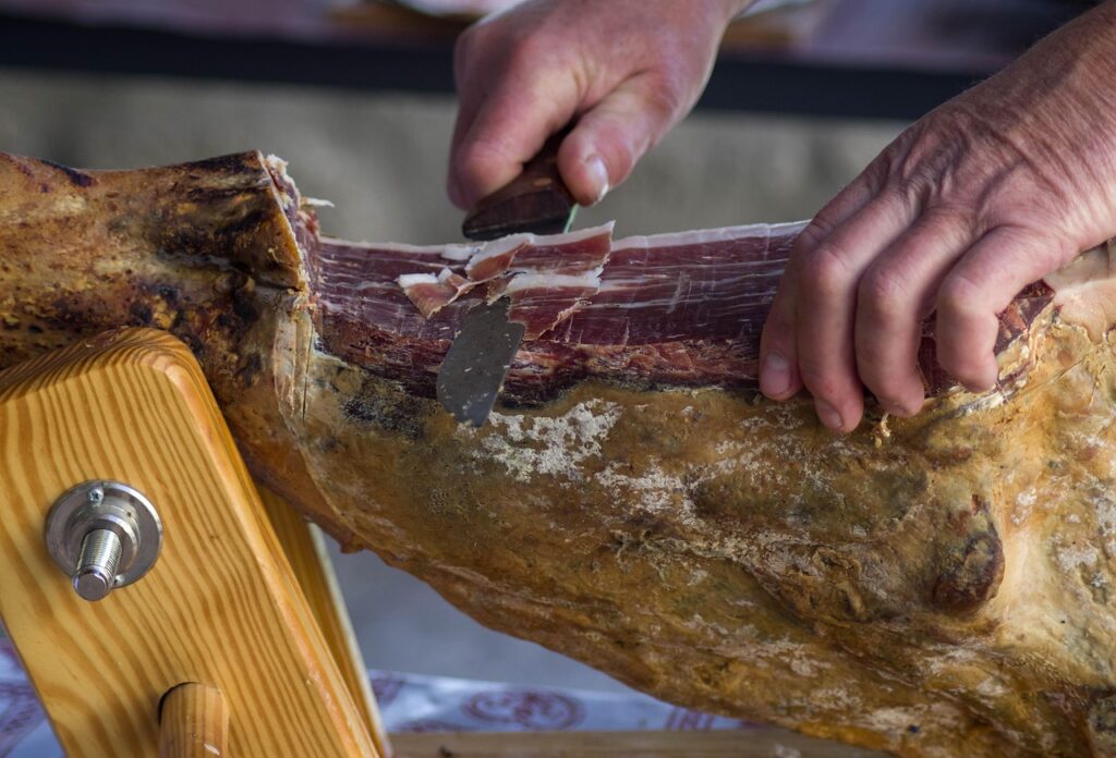 Hands slicing Prosciutto di Parma on a wooden ham stand.