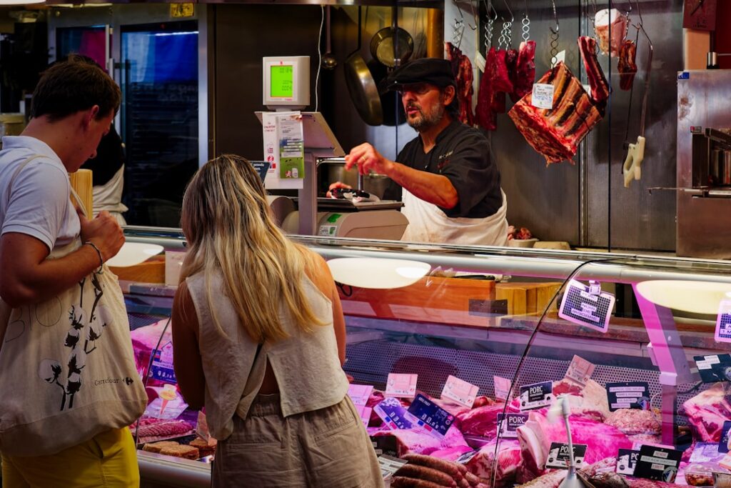 Butcher behind counter serving customers with hanging cured meats in Parma market.