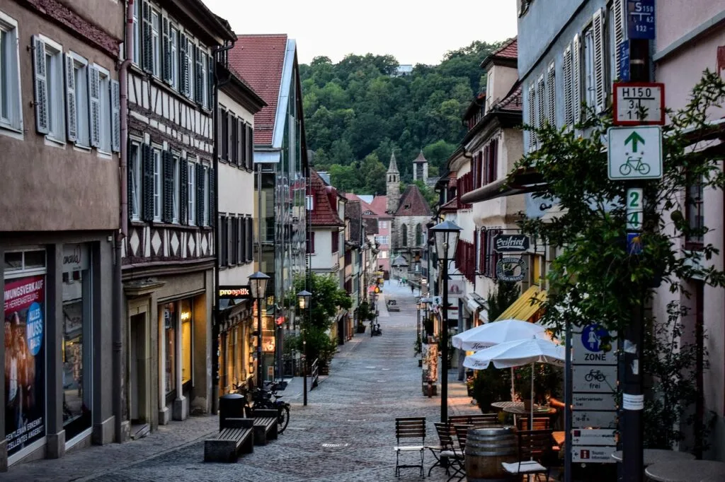 Rue pavée piétonne bordée de boutiques et terrasses, vue sur collines