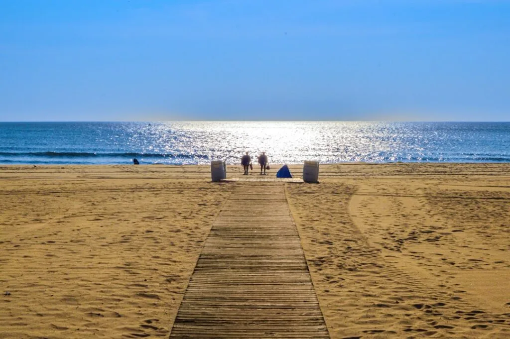 Passerelle en bois vers la plage, mer scintillante et deux promeneurs à Grado