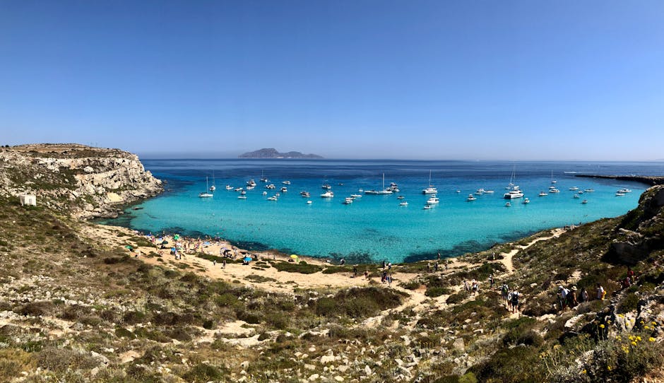 Baia di acque turchesi con numerose barche e spiaggia rocciosa in Sicilia
