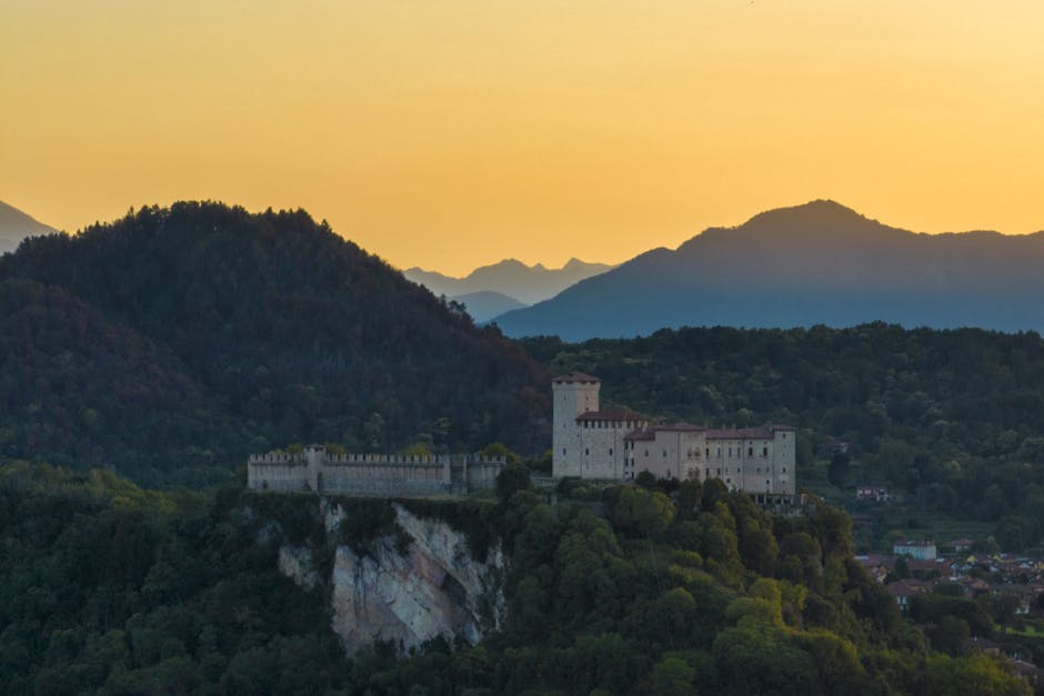 Medieval hilltop castle surrounded by forested hills under golden sunset.