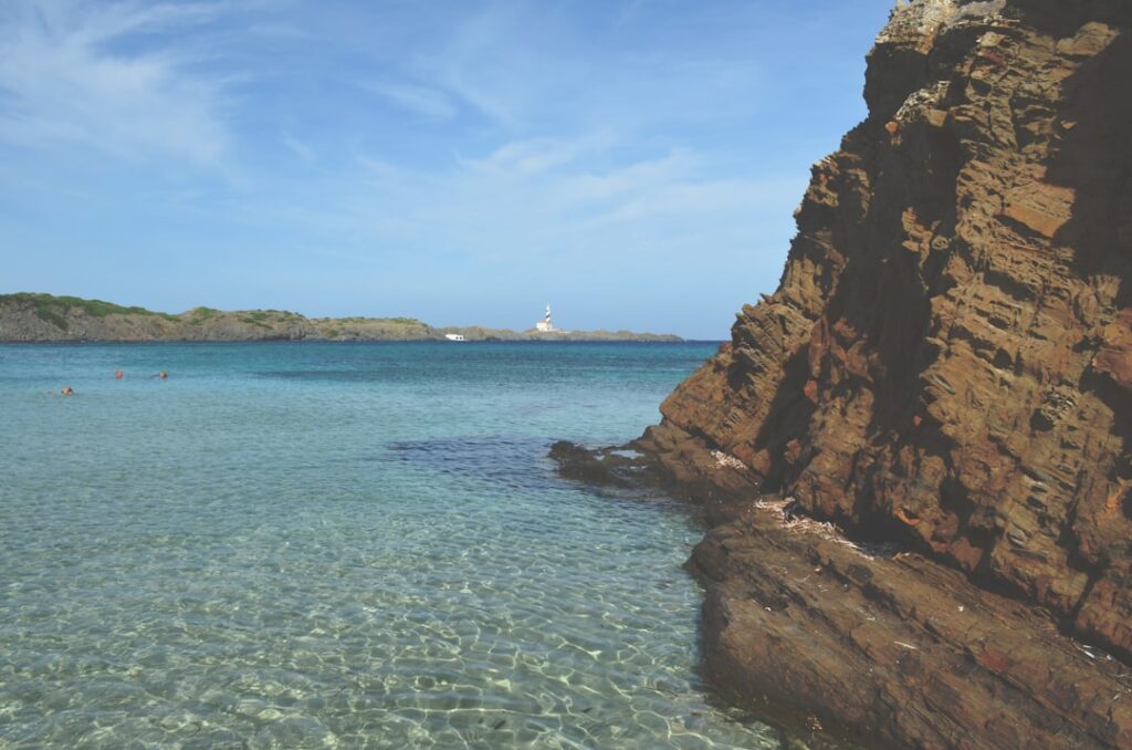 Kraakhelder turquoise water bij rotsachtige kust met vuurtoren op afstand