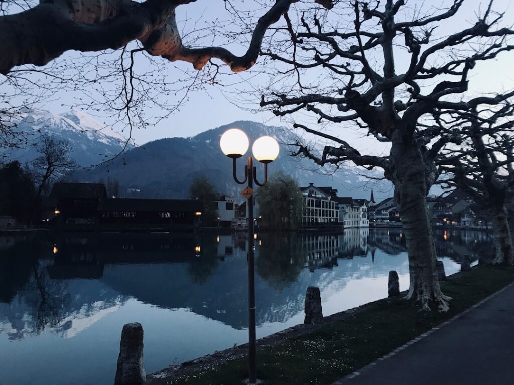 Uferpromenade mit Laterne und spiegelnder Bergkulisse nahe Trient am Fluss.