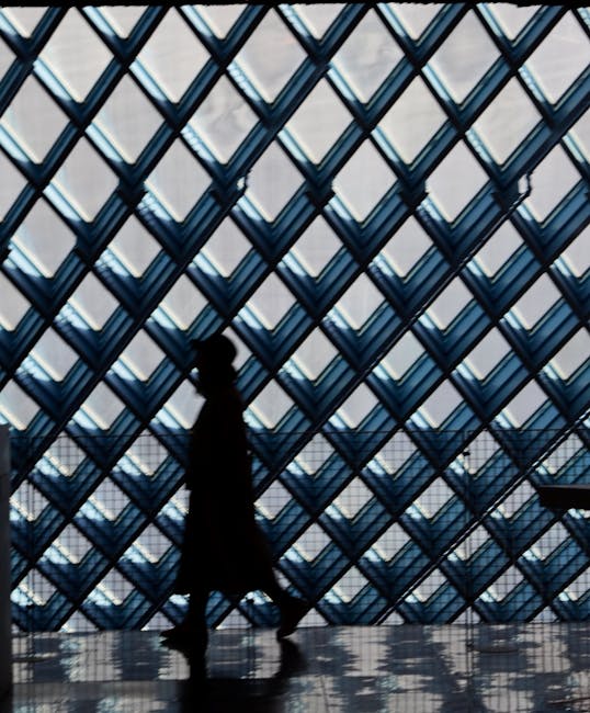 Silhouette einer gehenden Person vor rautenförmiger Glasfassade, Boden reflektiert