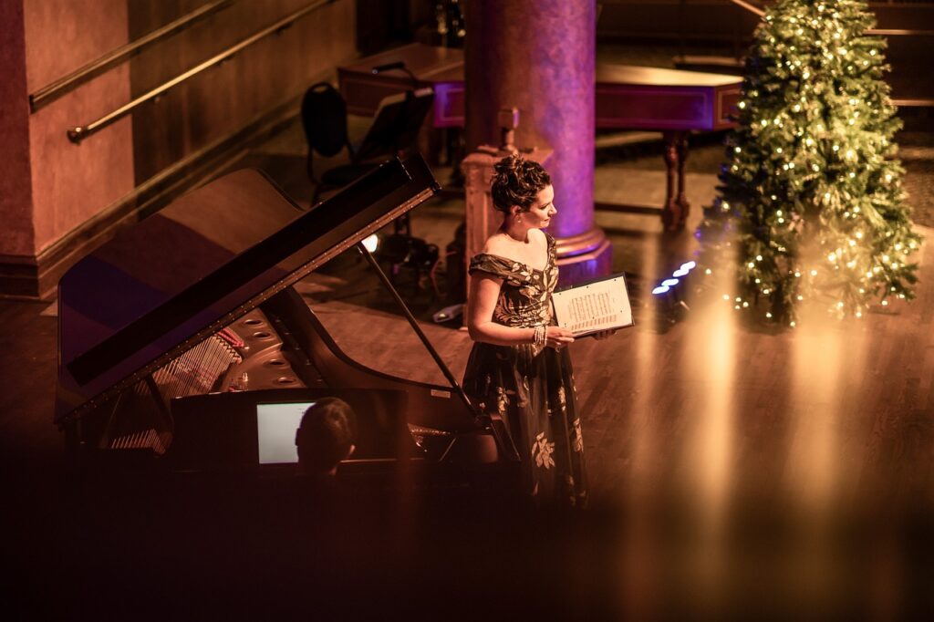 Soprano performing beside a grand piano in a warmly lit hall.