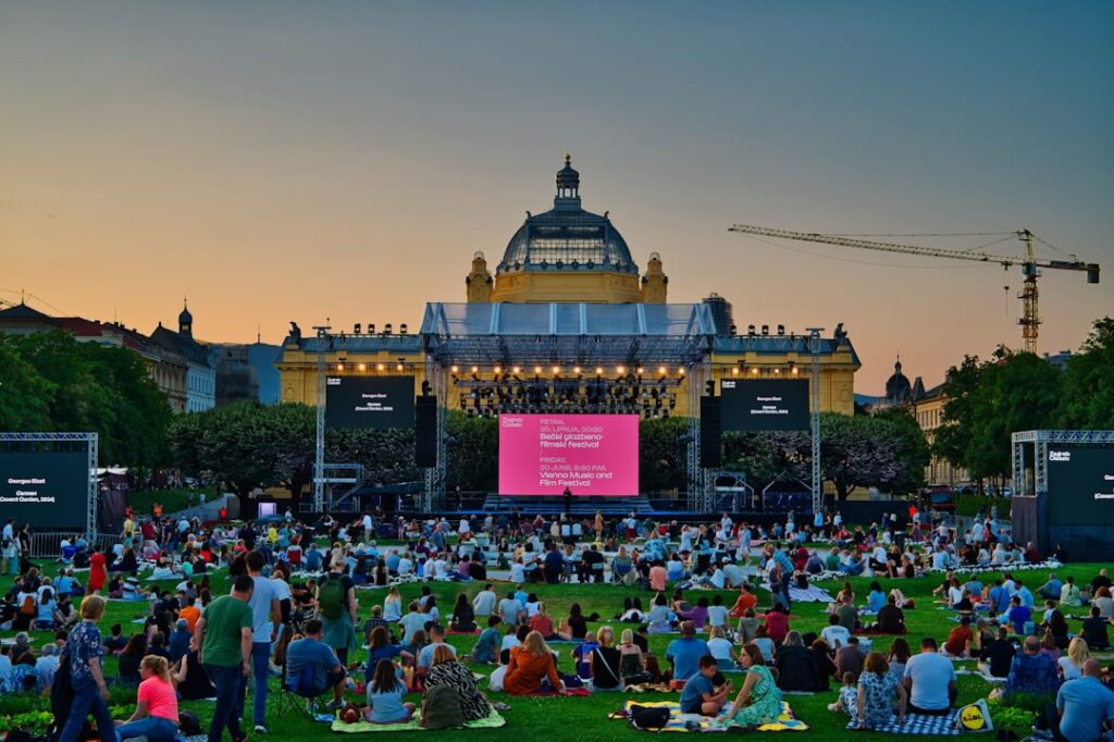 Crowds picnic on lawn facing an illuminated outdoor concert stage at dusk.