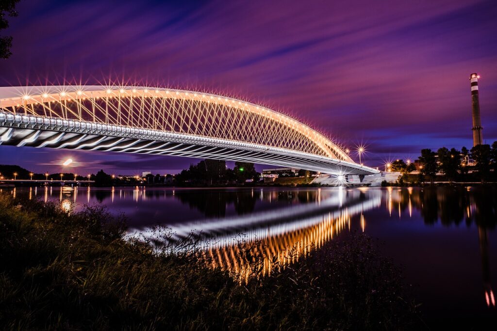 Pont moderne illuminé reflété dans l'eau sous un ciel violet nocturne.