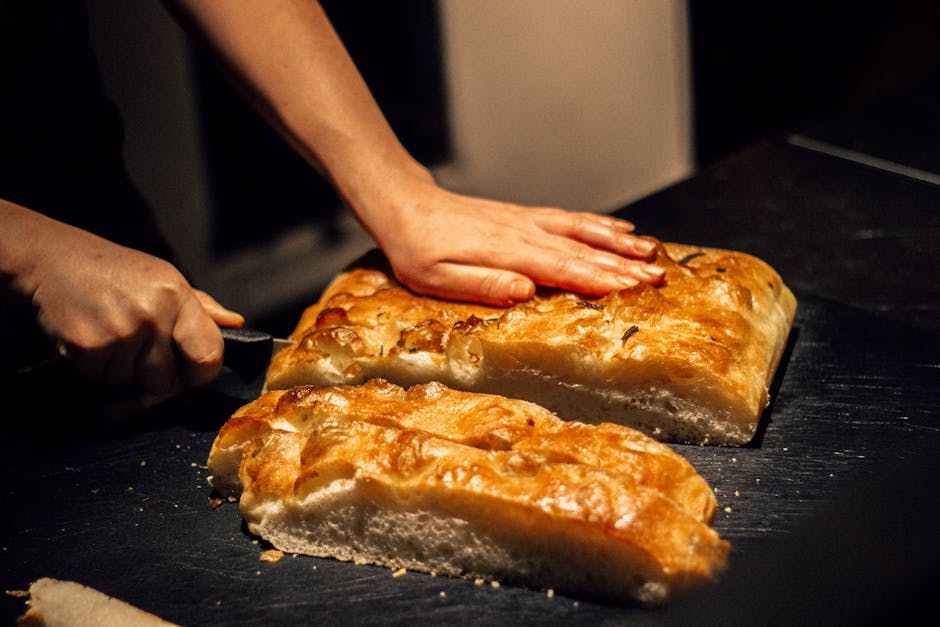 Hands slicing golden sourdough focaccia loaf on dark counter in Bari