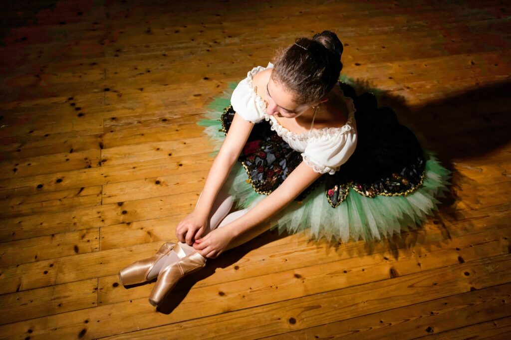 Ballerina in tutu tying pointe shoes on a wooden stage floor