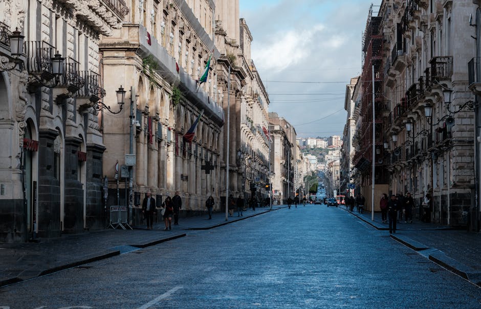 Wide historic street in central Catania lined with baroque buildings and flags