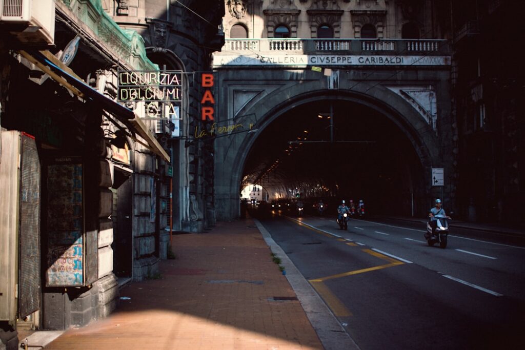 Sunlit Catania street leading to arched tunnel with neon 'BAR' sign.