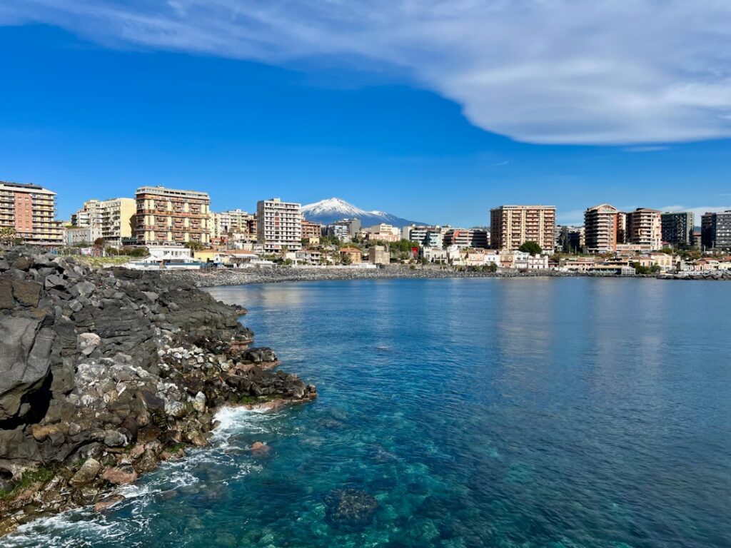 Litorale roccioso con skyline di Catania e l'Etna innevato sullo sfondo