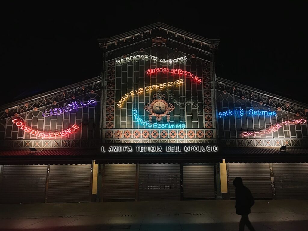 Neon-lit L'Antica Tettoia dell'Orologio market facade in Piedmont at night, pedestrian silhouette