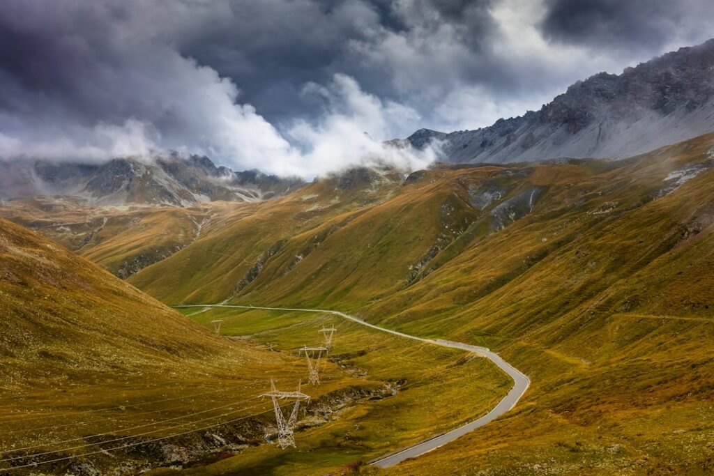 Gewundene Passstraße mit Strommasten in goldenem Tal unter Wolken im Piemont