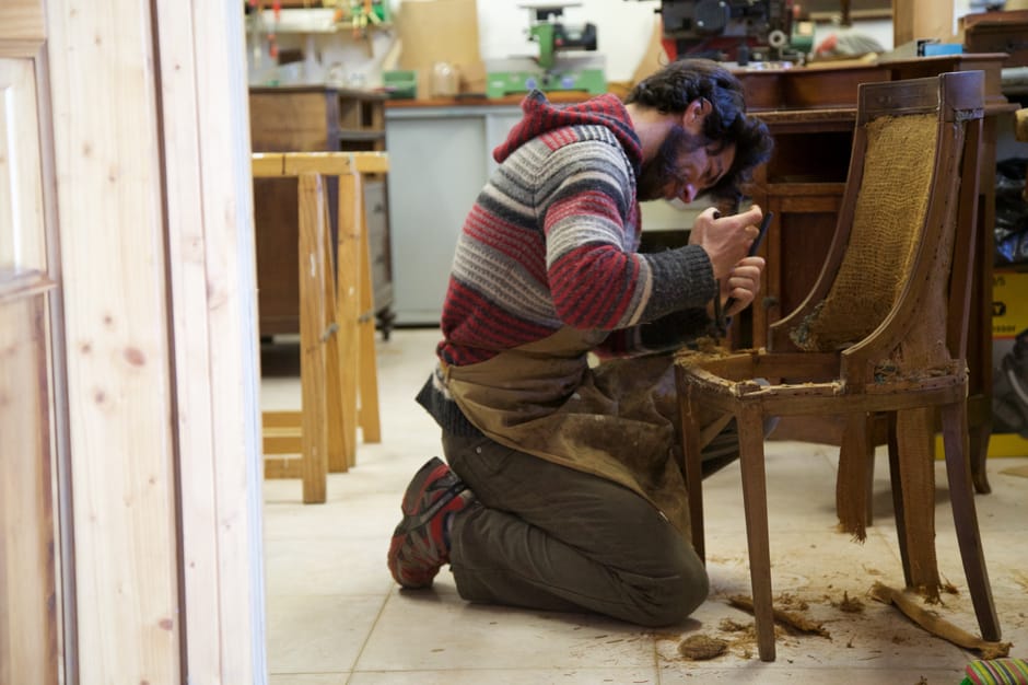 Artisan agenouillé rénovant une chaise en bois dans un atelier piémontais
