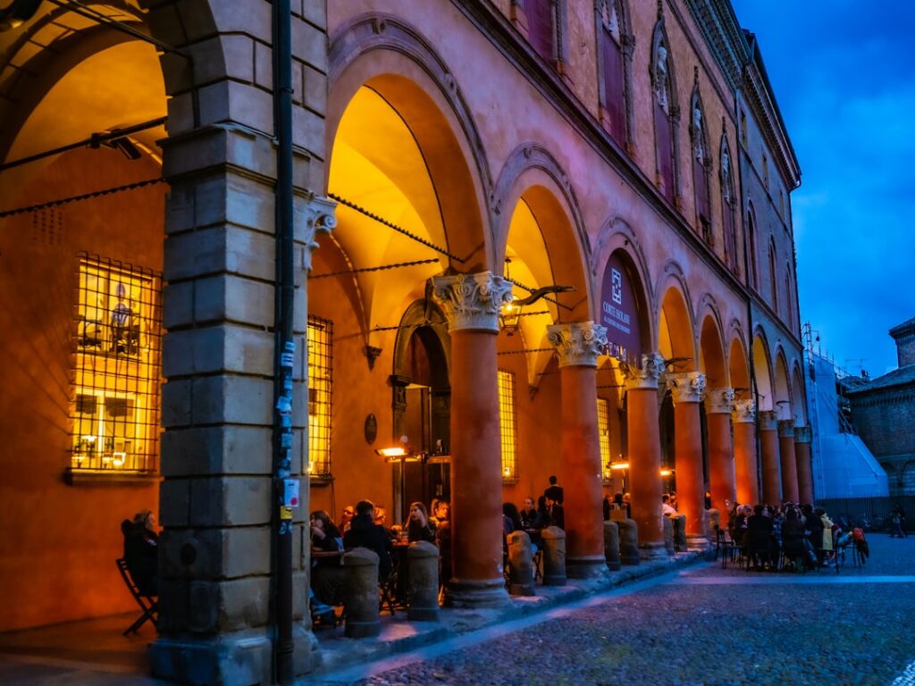 Arcades éclairées d'une place italienne au crépuscule, terrasses et passants