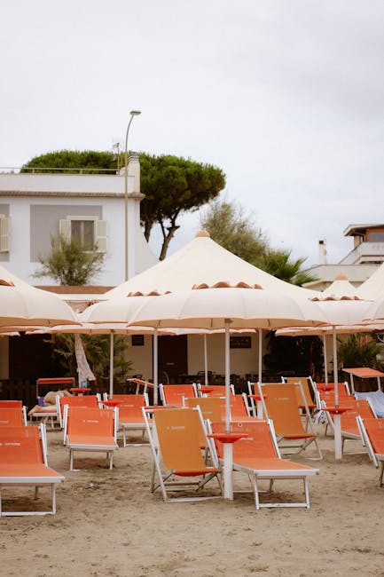 Leere orange Liegestühle und beige Sonnenschirme vor Häusern in Lido di Ostia, Latium