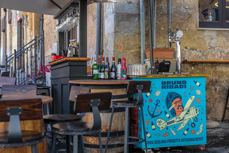 Street bar cart with Bruno Ribadi artwork and stools against stone wall in Latium alleyway