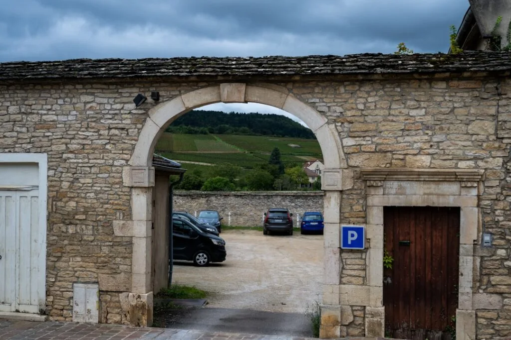 Portail en pierre ouvrant sur un parking et des vignes lointaines