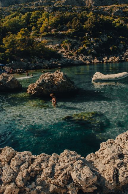 Person wading in shallow turquoise cove with boat near Rhodes shore