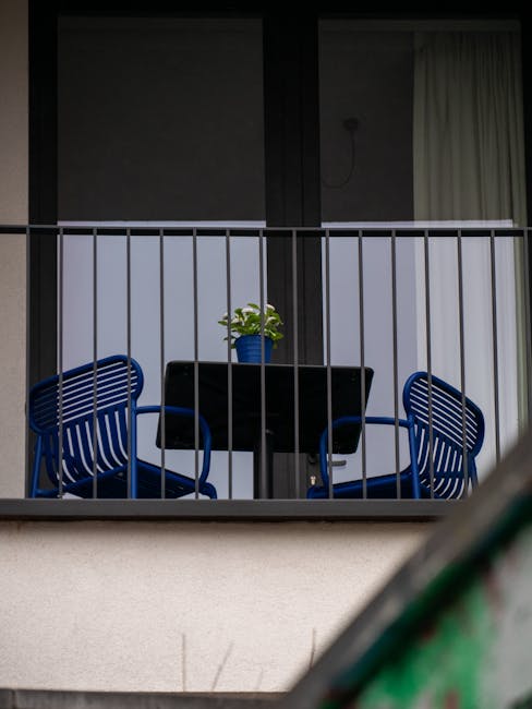 Small balcony with blue chairs and potted plant behind metal railing.
