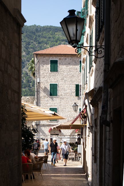Sunlit narrow alley in Kotor opening onto cafés and walking visitors.
