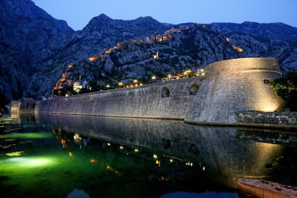 Illuminated Kotor city walls reflected in calm moat at dusk