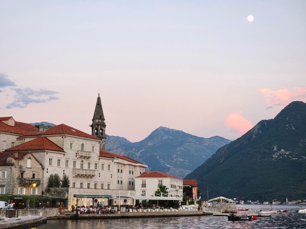 Kotor waterfront at dusk with red roofs and mountain backdrop.