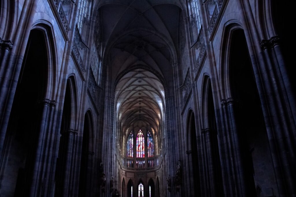Dark cathedral nave with tall arches and colorful stained glass.