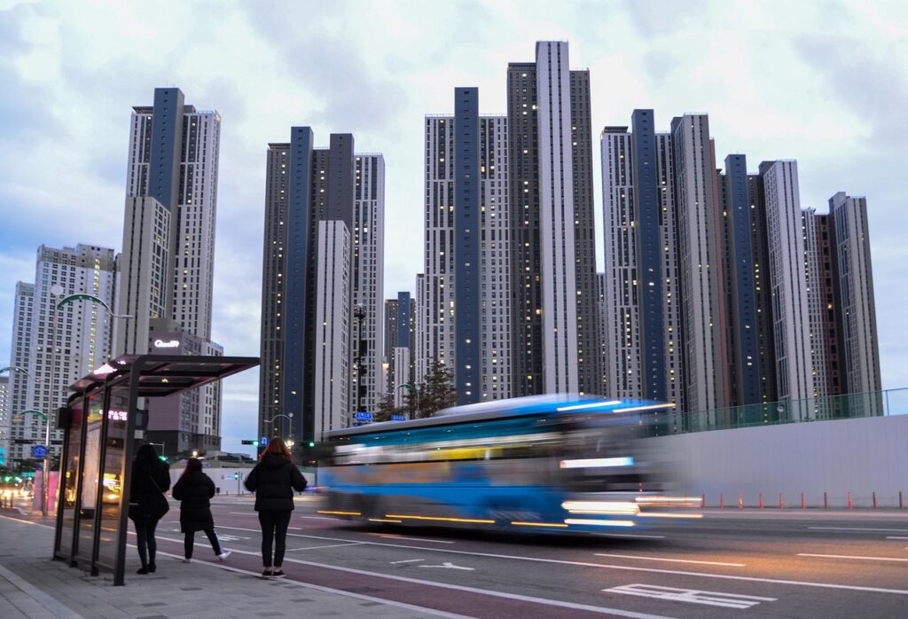 Blurred blue bus speeding past commuters at urban bus stop with high-rise towers