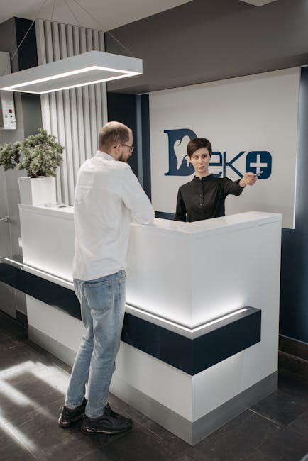 Clinic receptionist gestures to a male patient at a white desk in Tirana