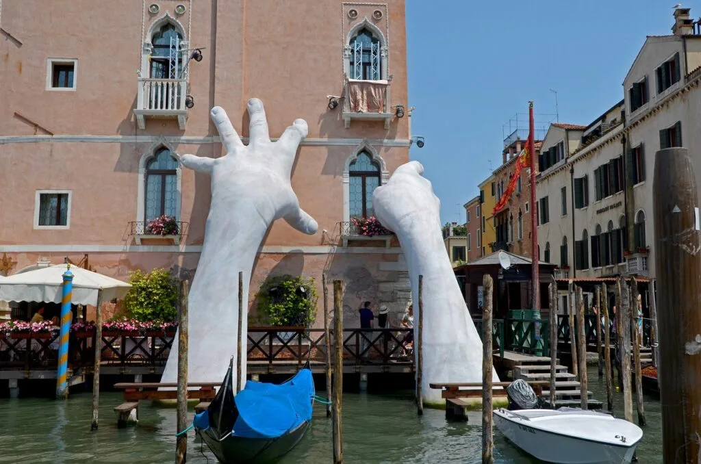 Deux grandes mains blanches émergeant de l'eau soutenant une façade vénitienne.