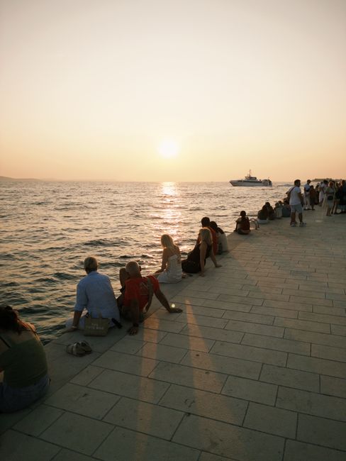 Passeggiata sul lungomare di Zara con panchine, il Sea Organ e persone al tramonto